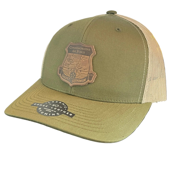 An olive green trucker cap with a tan mesh back and a brown embroidered shield patch on the front.